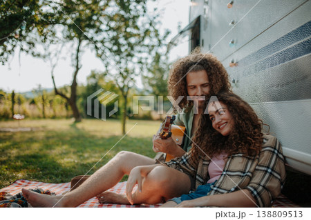 Couple sitting by caravan, enjoying peaceful moment. Man playing guitar and singing song to his girlfriend. Couple sitting by caravan, enjoying peaceful moment. Man playing guitar and singing song to his girlfriend. 138809513