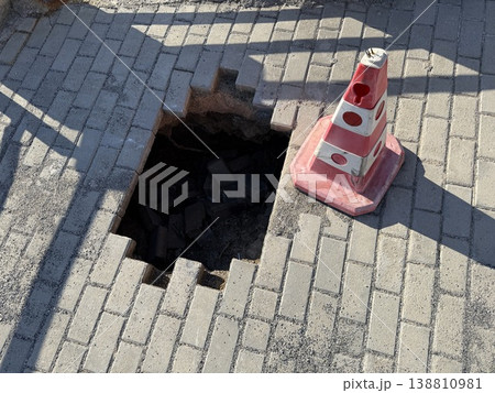 Damaged road surface with missing brick pavers and a warning cone nearby under sunlight Damaged road surface with missing brick pavers and a warning cone nearby under sunlight 138810981