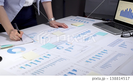 Business professional analyzing financial data with charts and graphs, working late in an office preparing reports and organizing information with sticky notes and a pen 138814517