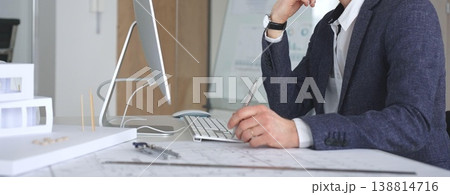 Man architect wearing glasses, is designing building plans on computer, thinking and concentrating on construction project blueprints at office desk. Business and architecture concept 138814716