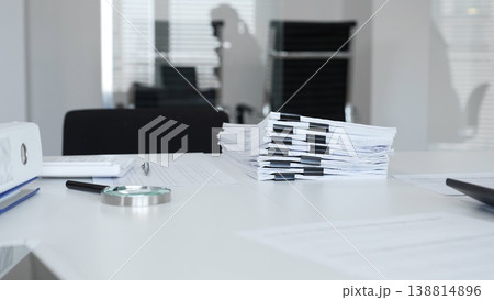 Office desk with stack of paper and magnifying glass, symbolizing heavy bureaucracy and business workload. Audit and taxes in business Office desk with stack of paper and magnifying glass, symbolizing heavy bureaucracy and business workload. Audit and taxes in business 138814896