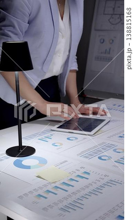 Professional businesswoman working at a desk surrounded by financial reports, charts, and graphs, focusing on data analysis to develop strategic plans and improve performance 138815168