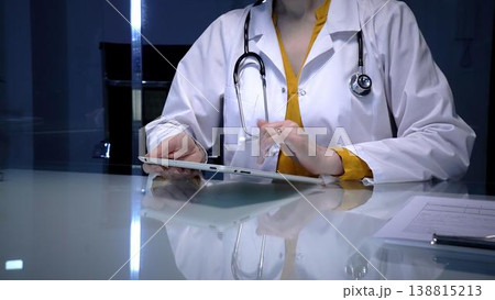 Female doctor wearing a white lab coat and stethoscope, using a digital tablet for telemedicine consultation, reviewing patient records, or managing hospital information system 138815213