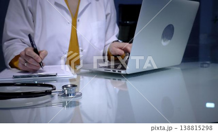 Stethoscope resting on a white reflective desk while medical professional working in clinic office. Wearing lab coat. Typing on laptop and writing notes. Stethoscope resting on a white reflective desk while medical professional working in clinic office. Wearing lab coat. Typing on laptop and writing notes. 138815298