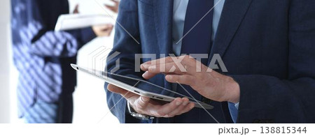 Businessman in a blue suit using a tablet during a corporate meeting, interacting with the touchscreen as blurred colleagues collaborate in the background, in office. Business people concept 138815344