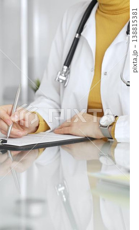Doctor wearing stethoscope and yellow sweater under white lab coat, making notes on a clipboard in a bright medical office. Medicine, healthcare and science concept 138815381