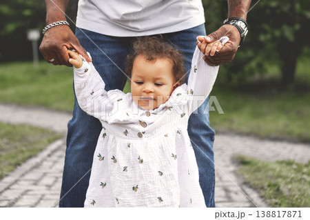 Baby girl taking first steps on park path, African American father's hands holding arms from behind, child milestone candid lifestyle outdoors 138817871