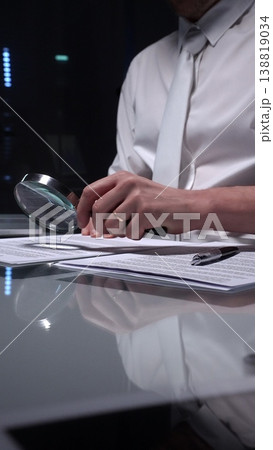 Person examining a document using a magnifying glass, focusing on fine print and terms in a legal agreement, working late. Audit and overworking concepts Person examining a document using a magnifying glass, focusing on fine print and terms in a legal agreement, working late. Audit and overworking concepts 138819034