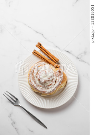 Freshly baked cinnamon roll with white icing on a plate and fork on a light marble background. Freshly baked cinnamon roll with white icing on a plate and fork on a light marble background. 138819051