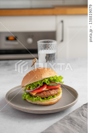 Beef burger with tomato, cheese, and lettuce on a plate on a kitchen table with glass of water Beef burger with tomato, cheese, and lettuce on a plate on a kitchen table with glass of water 138819079