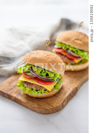 Two Beef burgers with tomato, cheese, and lettuce on a wooden board on a white marble table Two Beef burgers with tomato, cheese, and lettuce on a wooden board on a white marble table 138819082