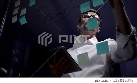 Man in eyeglasses organizing thoughts and ideas for business optimization on a clear glass board. Creating a strategic roadmap for innovation and project planning Man in eyeglasses organizing thoughts and ideas for business optimization on a clear glass board. Creating a strategic roadmap for innovation and project planning 138819379