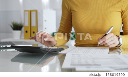 Businesswoman wearing yellow casual sweater is calculating budget and reviewing financial documents using a laptop and calculator for home finances. Audit and taxes in business Businesswoman wearing yellow casual sweater is calculating budget and reviewing financial documents using a laptop and calculator for home finances. Audit and taxes in business 138819390