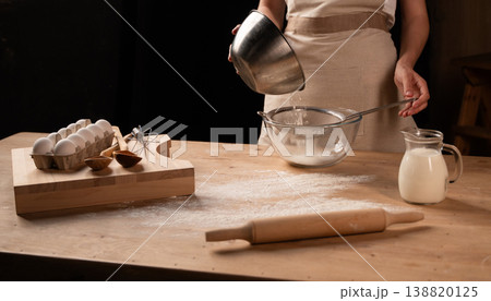 Woman sifting flour over a glass bowl, surrounded by baking ingredients like eggs, milk, and a rolling pin Woman sifting flour over a glass bowl, surrounded by baking ingredients like eggs, milk, and a rolling pin 138820125