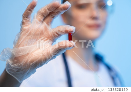 Healthcare professional wearing protective glove holding a small red capsule. Concept for medication, research, and illness prevention Healthcare professional wearing protective glove holding a small red capsule. Concept for medication, research, and illness prevention 138820139