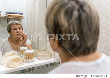Senior woman doing a skincare routine, cleaning her face with a cotton pad in front of a mirror, focusing on self-care. Real people, life of an elderly person, daily habits routine Senior woman doing a skincare routine, cleaning her face with a cotton pad in front of a mirror, focusing on self-care. Real people, life of an elderly person, daily habits routine 138820171