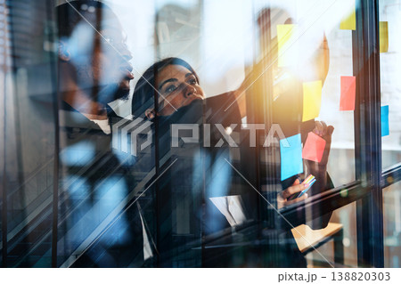 Team members collaborate at an office meeting while discussing ideas on a glass wall with colorful sticky notes in view Team members collaborate at an office meeting while discussing ideas on a glass wall with colorful sticky notes in view 138820303