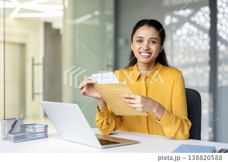 Businesswoman receiving good news from mail, opening a manila envelope with important documents at a desk in a modern office while smiling at the camera 138820588