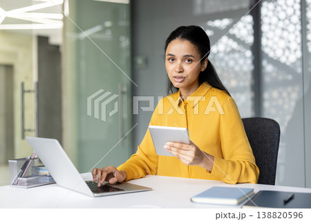 Young professional woman sitting at her desk, working efficiently with a laptop and holding a digital tablet in a modern office environment, demonstrating productivity and business connectivity 138820596