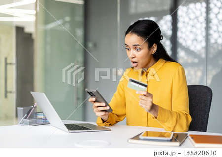 Biracial woman at desk looking shocked at her smartphone while holding a credit card, laptop open, reacting to unexpected online banking charges or potential fraud notification Biracial woman at desk looking shocked at her smartphone while holding a credit card, laptop open, reacting to unexpected online banking charges or potential fraud notification 138820610