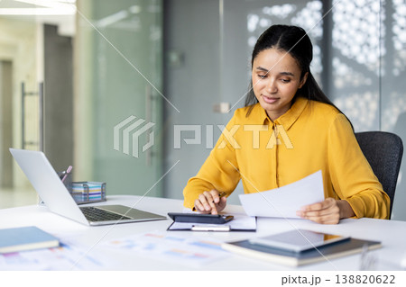 Young professional woman or accountant sitting at an office desk, analyzing financial documents and using a calculator while working with a laptop and other paperwork, managing business finances Young professional woman or accountant sitting at an office desk, analyzing financial documents and using a calculator while working with a laptop and other paperwork, managing business finances 138820622
