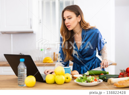 Young woman in blue robe cooking and watching TV series on laptop Young woman in blue robe cooking and watching TV series on laptop 138822428
