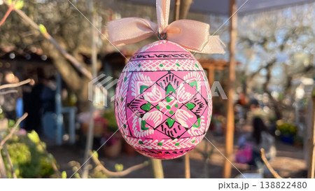 Traditional painted Easter egg with white ribbon hanging on budding tree branch in sunny garden 138822480