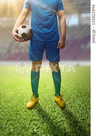 Professional soccer player in a blue uniform holding a soccer ball while standing on a lush green field inside a bright stadium Professional soccer player in a blue uniform holding a soccer ball while standing on a lush green field inside a bright stadium 138822949