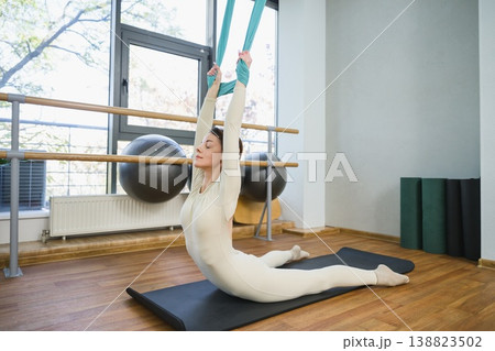 Young woman sitting on mat performing stretch with aerial yoga hammock. Young woman sitting on mat performing stretch with aerial yoga hammock. 138823502