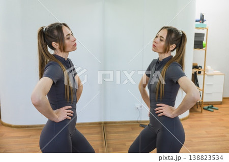 Young woman standing in fitness studio looking at her reflection in mirror. Young woman standing in fitness studio looking at her reflection in mirror. 138823534