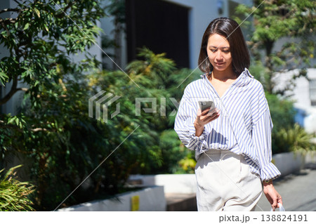 買い出しの帰り道、スマホを片手に笑顔で休日の散歩を楽しむ日本人女性。 買い出しの帰り道、スマホを片手に笑顔で休日の散歩を楽しむ日本人女性。 138824191