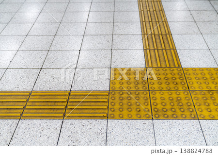 Yellow tactile paving tiles on a transit station floor for visually impaired guidance. Braille blocks with bumps and stripes on light stone tiles for safety and accessibility in public urban spaces. 138824788