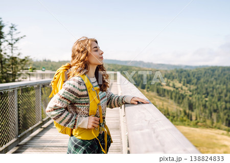 Happy woman with yellow backpack on wooden pedestrian bridge enjoying mountain scenery. 138824833