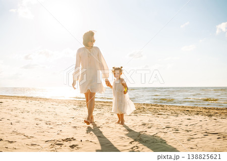 A mother and daughter enjoy a sunny beach stroll, as waves gently lap at their feet during sunset 138825621