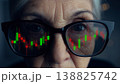 A person looks at stock market data reflected in their thick rimmed glasses. The setting shows trading activity. Bright colors represent market trends. 138825742