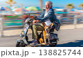 A man moves quickly on a mobility scooter along a boardwalk. A dog sits in front, both smiling amidst beach umbrellas and people enjoying the day. 138825744