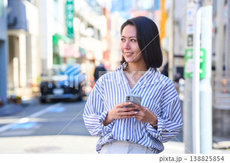 買い出しの途中にスマホを見て微笑む日本人女性。休日の街角での散歩。 買い出しの途中にスマホを見て微笑む日本人女性。休日の街角での散歩。 138825854