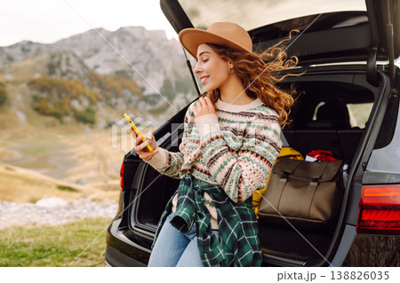 Young traveler woman sits on the rear of her car, smiling and using phone in scenic mountainous area Young traveler woman sits on the rear of her car, smiling and using phone in scenic mountainous area 138826035