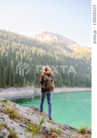 Back view young woman with backpack looking the view of the mountain lake in sunny weather. Back view young woman with backpack looking the view of the mountain lake in sunny weather. 138826223