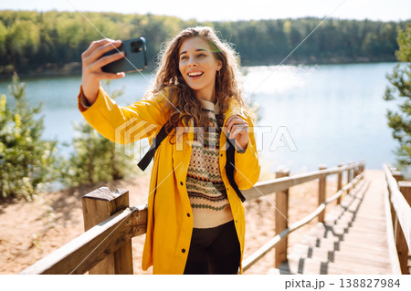 Happy woman with phone in hands taking selfie in forest. Young tourist enjoying spring day near lake 138827984