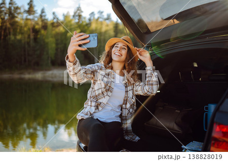 Selfie in nature. Happy woman sits in trunk of car, uses smartphone. Concept of traveling, blogging. 138828019