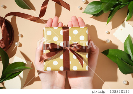 Hands holding a polka dot gift box with a brown ribbon over a beige background with leaves and pearls. Hands holding a polka dot gift box with a brown ribbon over a beige background with leaves and pearls. 138828133