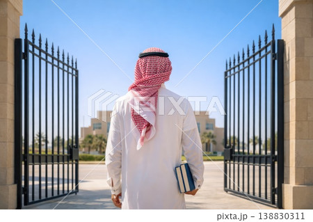 Middle Eastern man entering modern campus gate holding books from behind 138830311