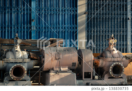 Bathed in the warm, low-angled light of late afternoon, a trio of heavy industrial pumps rests in quiet repose against a backdrop of weathered blue accordion gates. The central mauve motor is flanked Bathed in the warm, low-angled light of late afternoon, a trio of heavy industrial pumps rests in quiet repose against a backdrop of weathered blue accordion gates. The central mauve motor is flanked 138830735