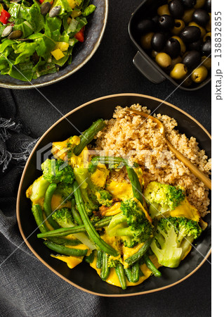 Healthy breakfast consists of a quinoa bowl with boiled eggs, green beans, and broccoli placed on a dark surface next to a cup of coffee and a woven cloth 138832855