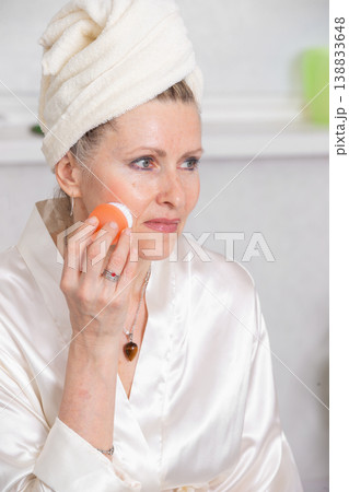Woman applying skincare treatment in a spa-like setting while wearing a silk robe and towel on her head Woman applying skincare treatment in a spa-like setting while wearing a silk robe and towel on her head 138833648