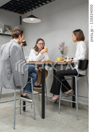 Three women gathered around high table in kitchen area chatting and eating during office lunch. Eating, chatting, kitchen, teamwork, lunch, office, women. 138834000
