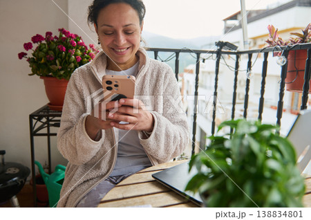 Smiling Woman Using Smartphone on Cozy Balcony Surrounded by Plants and Laptop Smiling Woman Using Smartphone on Cozy Balcony Surrounded by Plants and Laptop 138834801