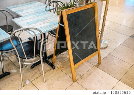 Empty blackboard (space for menu) on tiled floor, tables and chairs near. Mockup to add your own text. Empty blackboard (space for menu) on tiled floor, tables and chairs near. Mockup to add your own text. 138835630