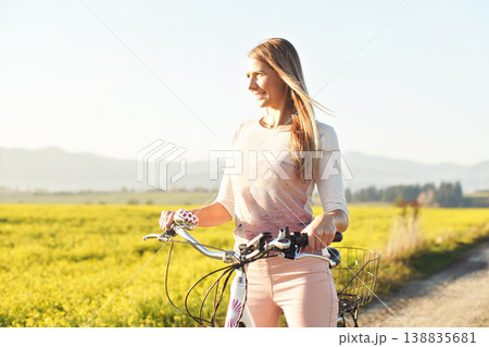 Young woman standing next to her bike over dusty country road, strong afternoon sun backlight in background shines on yellow flowers field Young woman standing next to her bike over dusty country road, strong afternoon sun backlight in background shines on yellow flowers field 138835681